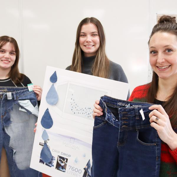 One students holds a pair of jeans with fabric cut out of it and another holds a poster.