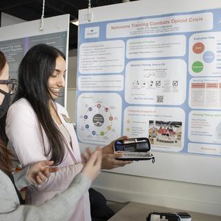 Two students discuss a poster behind them, one holds a naloxone kit