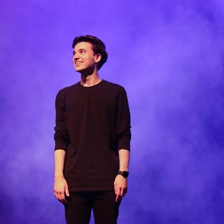 Alex Bhargava stands on the Malaspina Theatre stage looking off to the left. He wears all black and blue and purple smoke swirls around him.