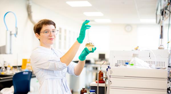 Misha Zvekic wears green gloves and holds a beaker with liquid in it in a chemistry lab.