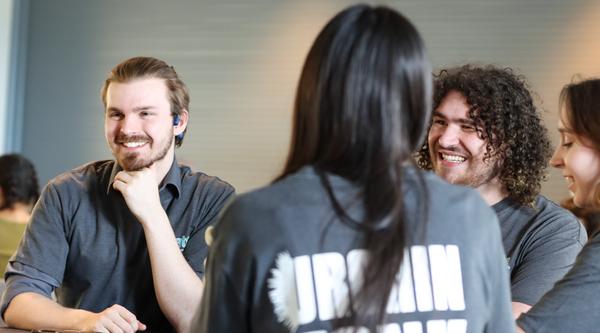 Three students wearing Urchin Tank T-Shirts talk while sitting at a table