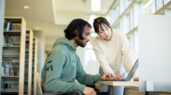 Two people look at a laptop screen in the library