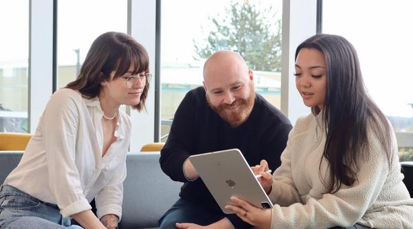 A woman, a man and another woman sitting on a chesterfield looking at a laptop together.