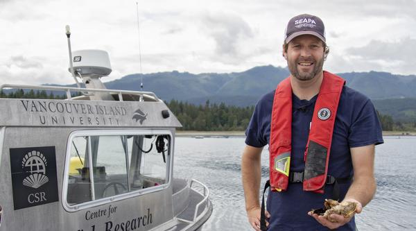 Dr. Timothy Green, VIU’s Canada Research Chair in Shellfish Health and Genomics stands on a dock next to a boat holding oysters in his hand.