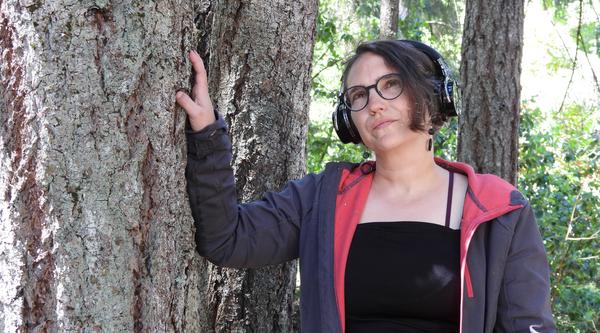 Sharon leans against a tree wearing headphones