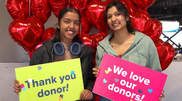 Two students hold up signs saying thank you donor and we love our donors and stand in front of red heart balloons