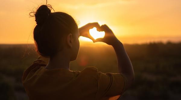Girl making a heart with her fingers and the sunset poking through