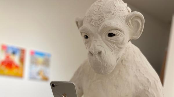 An ape made of white plaster and clay holds a cell phone, which it stares at intensely