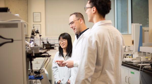 Dr. Kyle Duncan stands in front of a mass spectrometer with two students