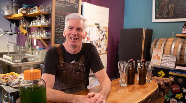 Dave Paul, owner of Love Shack Libations, leans against a wooden counter. Beer bottles and a glass is on his right.