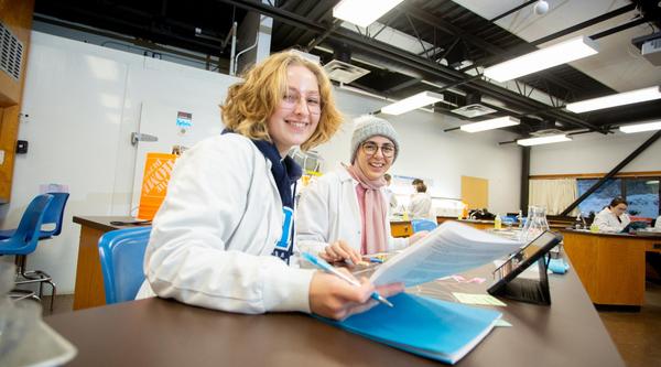 Two students look at a sheet in a lab, one has a pen in her hand, they are both smiling at the camera