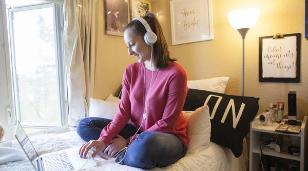 Student on a bed with headphones looking at a laptop