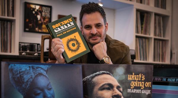 Paul Watkins holds up a copy of his new book in his right hand while resting his chin on the other.