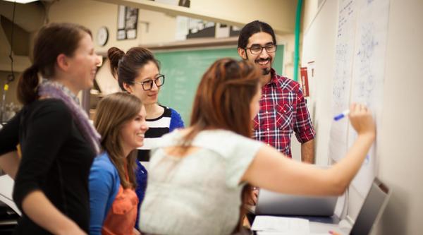 A group of students gathers around a student writing on a chalkboard