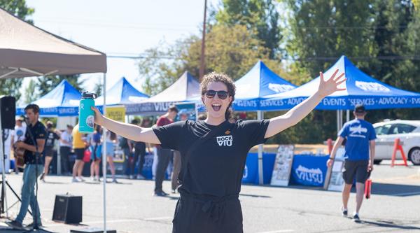 Student wearing a RockVIU T-Shirt stands with her arms stretched wide in front of tents and a performer playing the guitar
