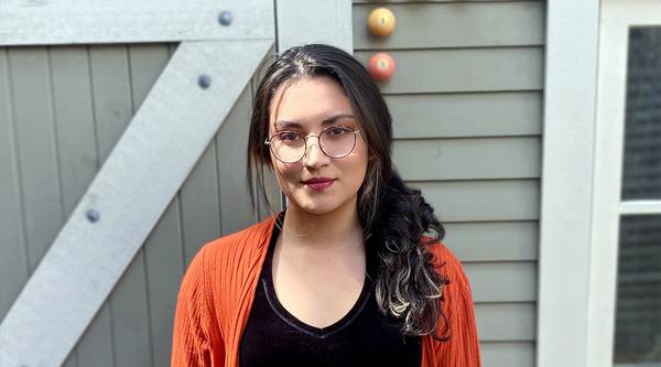 Sara Guzman stands in front of a building, wearing an orange sweater.