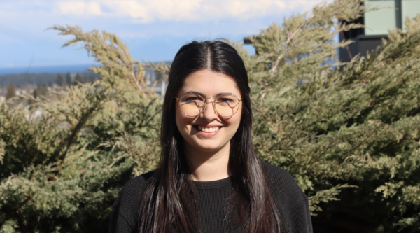 Mikaila Tombe standing outside in front of some bushes on a clear blue day smiling at the camera