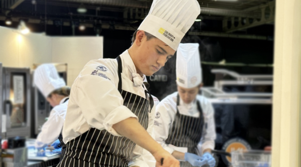 Ottis Crabbe wearing chef's hat and full cooking attire preparing a dish in a pot on a stove in an industrial kitchen during a competition.