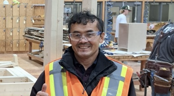 Ezat Haidary wering a reflective vests and safety goggles in the carpentry shop, giving a thumbs up and smiling at the camera.