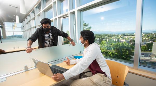 Two students in library wearing masks and talking with laptop between them