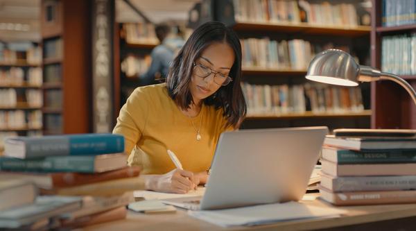A woman sits at a desk in a library with a laptop open surrounded by books