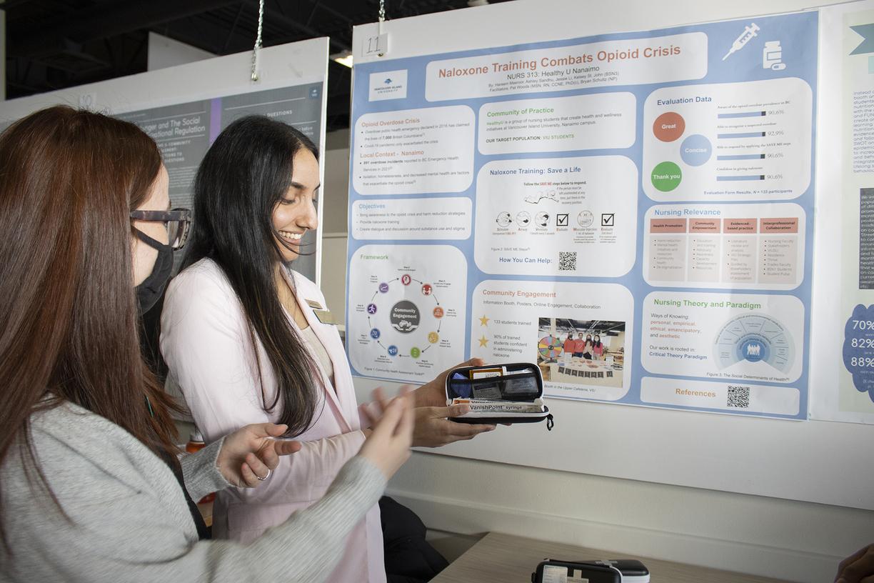 Two students discuss a poster behind them, one holds a naloxone kit