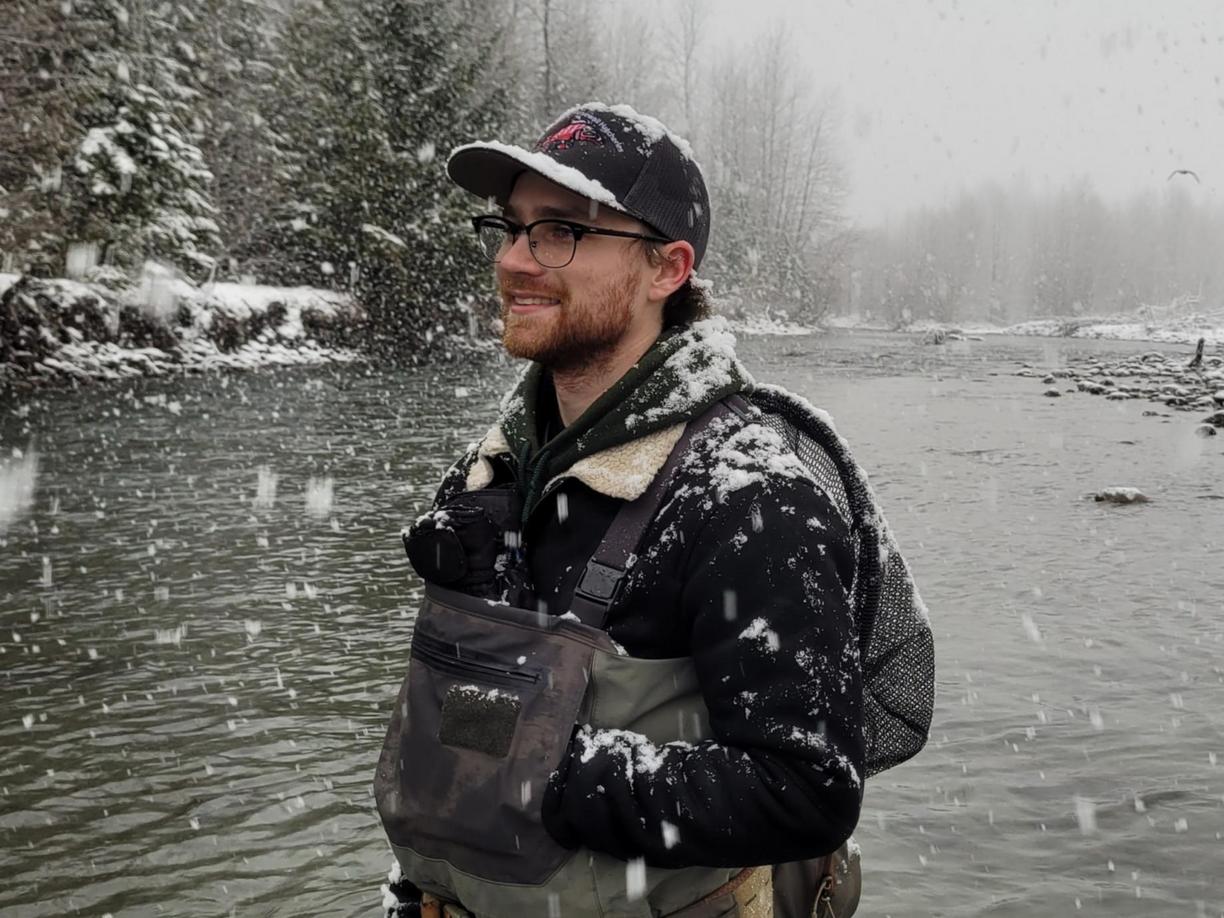 A young man clad in hipwaders is standing in a river with snowflakes falling around him