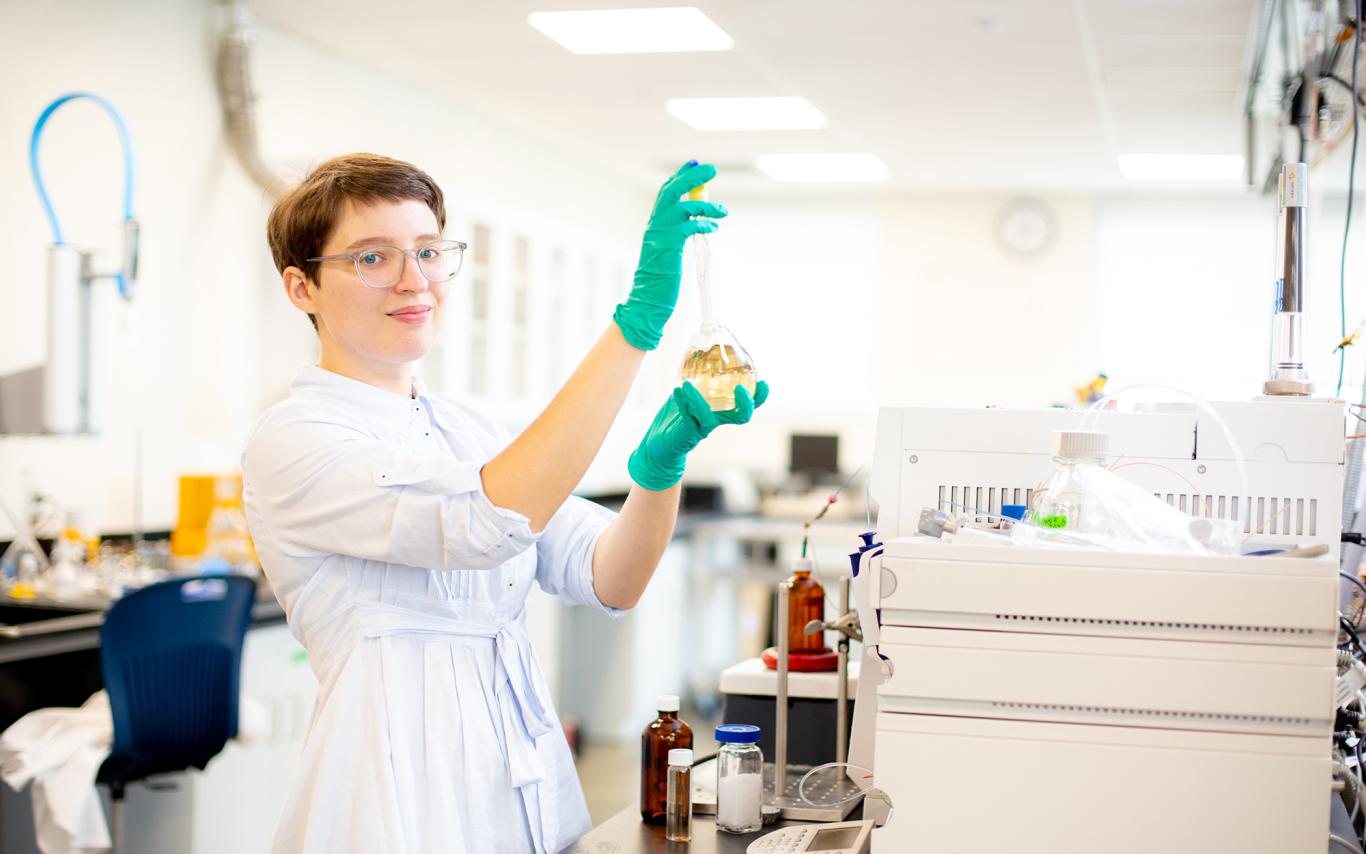 Misha Zvekic wears green gloves and holds a beaker with liquid in it in a chemistry lab.
