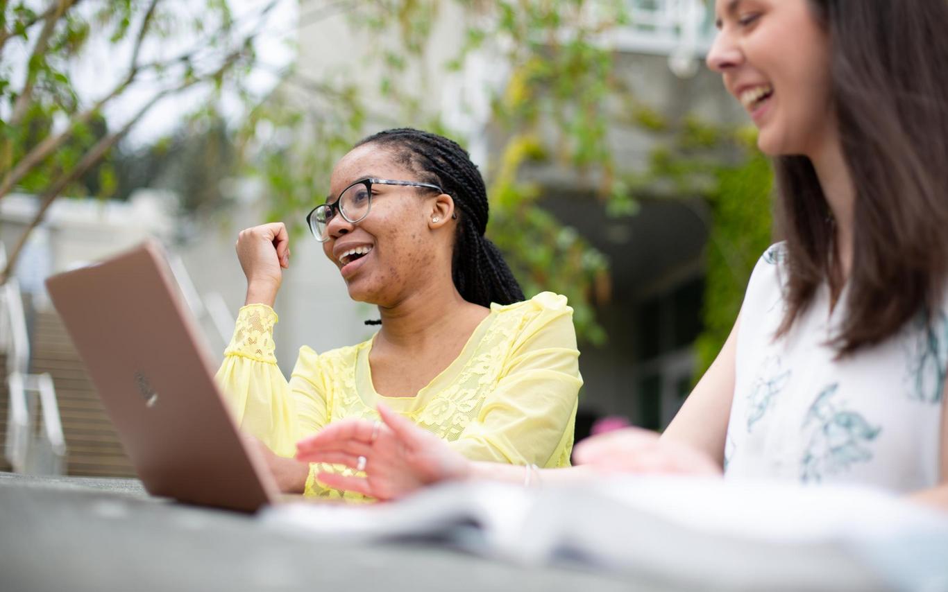 Two students sit at a table outdoors, one has a laptop open and the other has a textbook open. They are laughing together