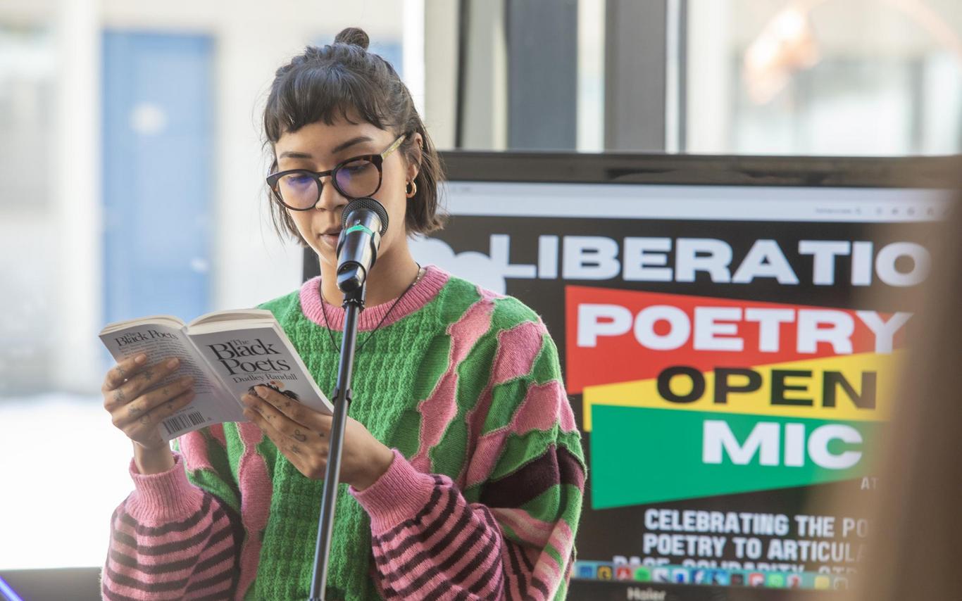 A woman wearing glasses and a pink and green sweater reads from a poetry book at a microphone