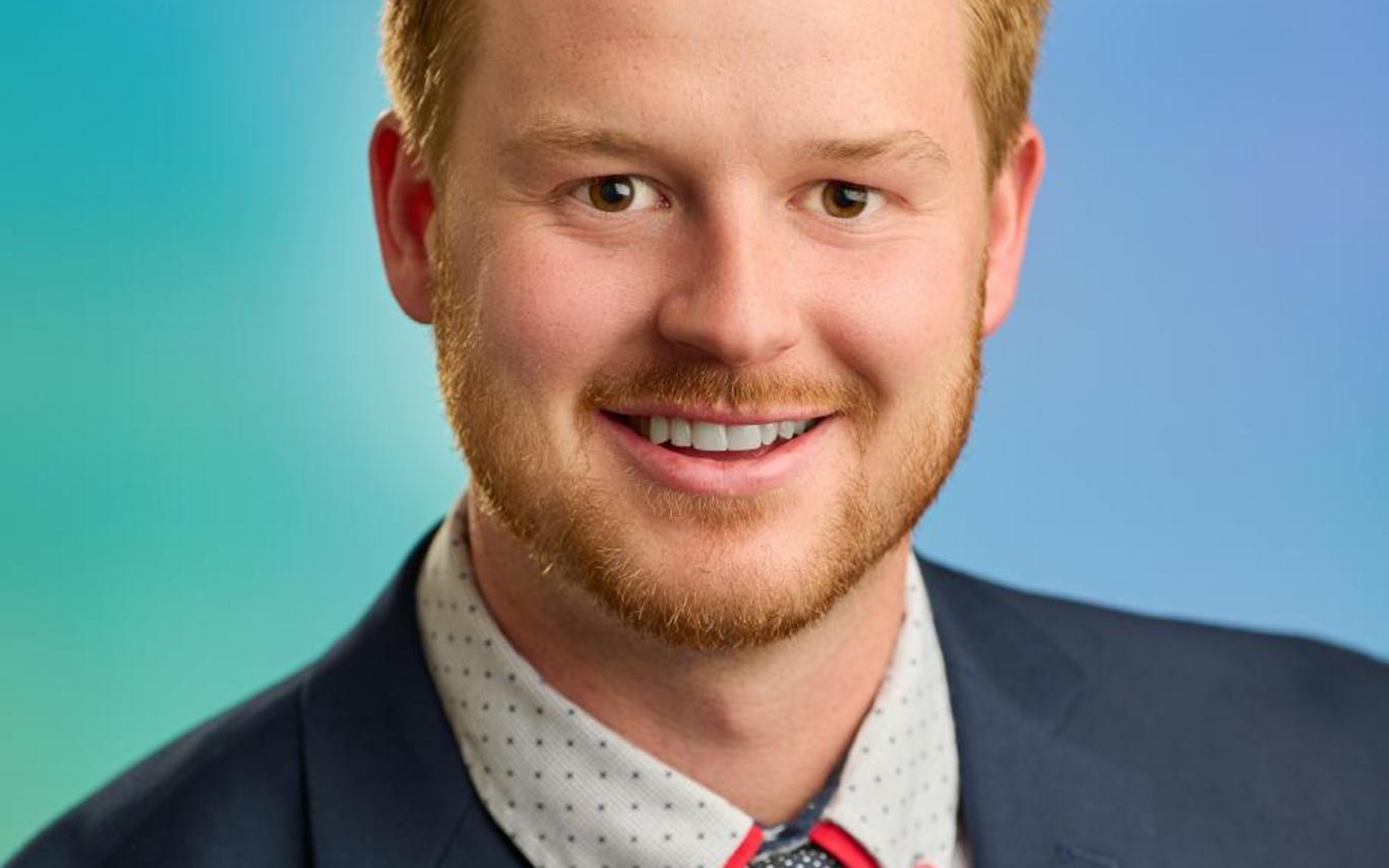 Mitch Turko wearing a suit and tie, smiling at the camera with a blue and green background.