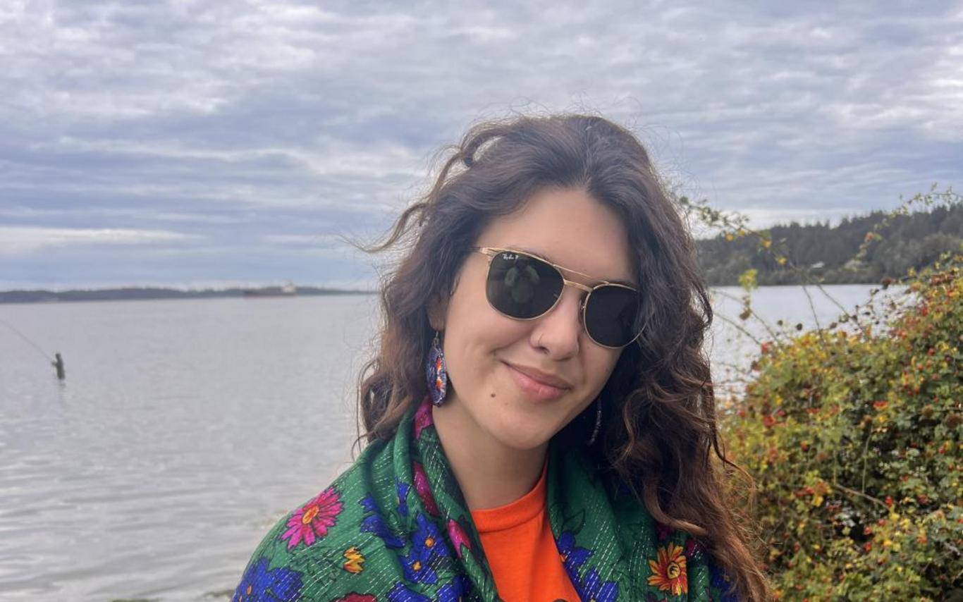 Alyona stands near a lake wearing a green shawl with pink, blue and yellow flowers over an Every Child Matters shirt.