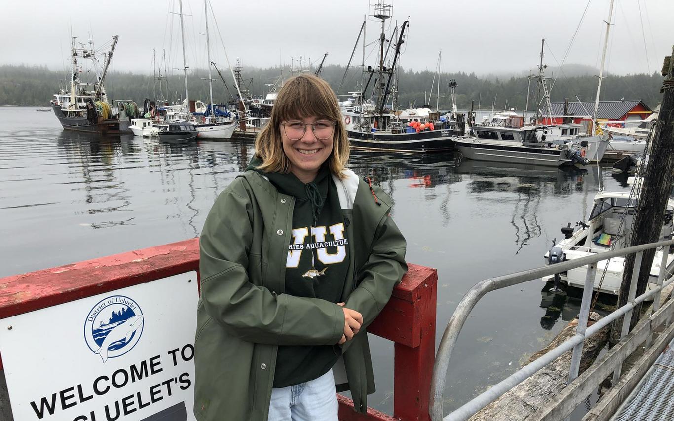 Allie Unger leans against a railing at Ucluelet harbour with boats anchored in the background.