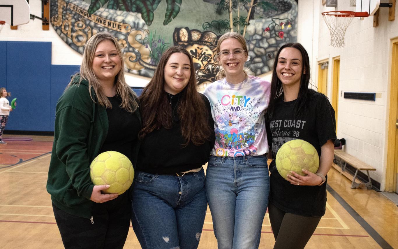 Four students in a gym holding volleyballs