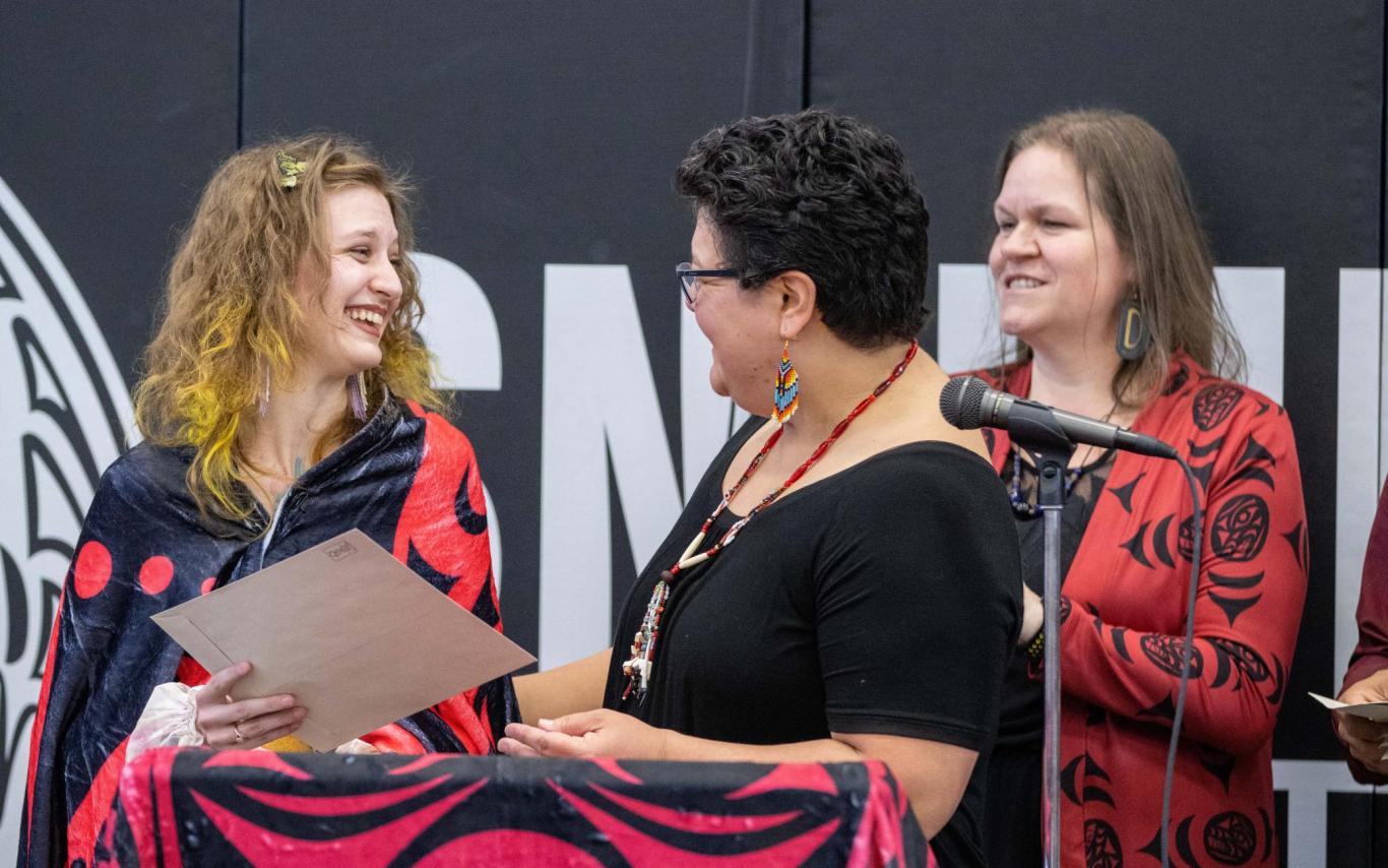 A student receives a folder from a woman, with another woman watching in the background