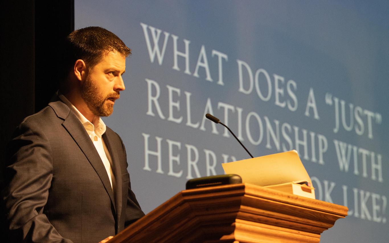 Man speaks at podium with text on a screen that reads: What does a just relationship with herring look like? 