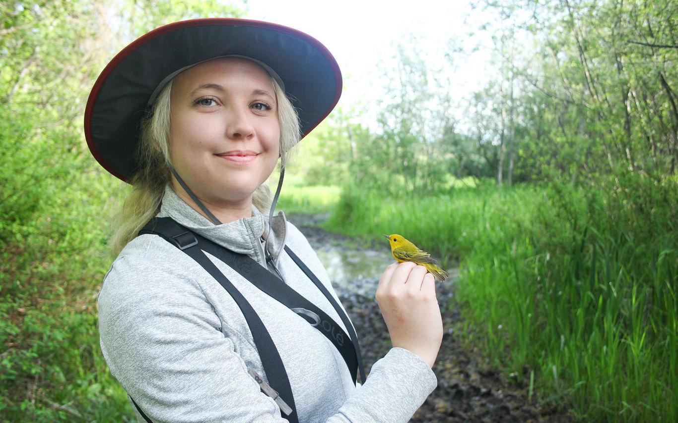 Chelsey Watts stands amongst bushes and trees holding a bird.