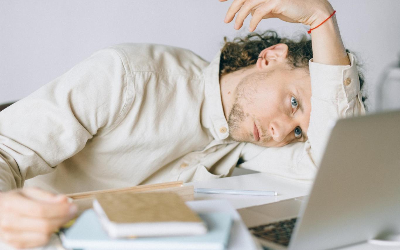 Student leaning on his arm staring dejectedly at a laptop screen with a pile of books beside him