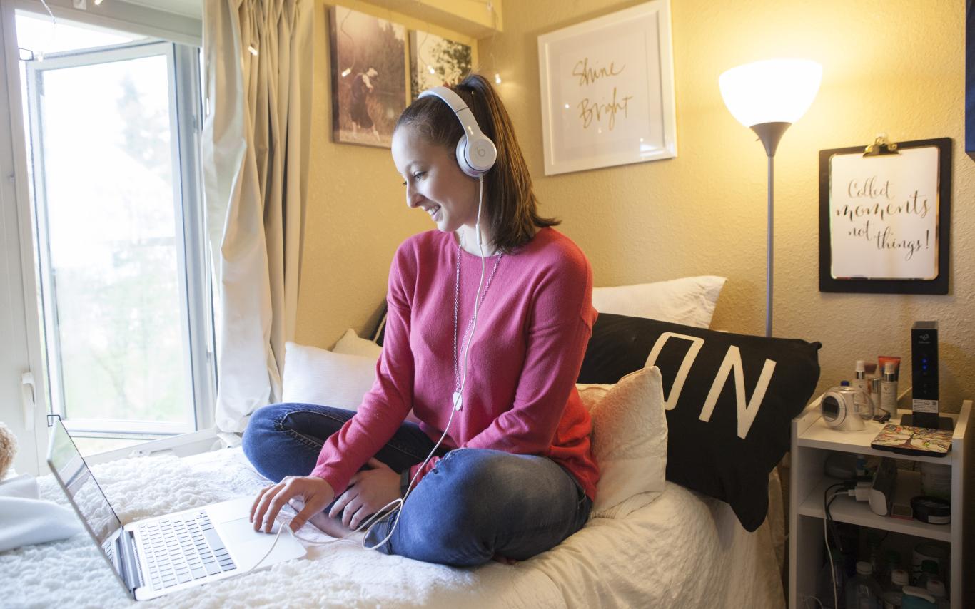 Student on a bed with headphones looking at a laptop