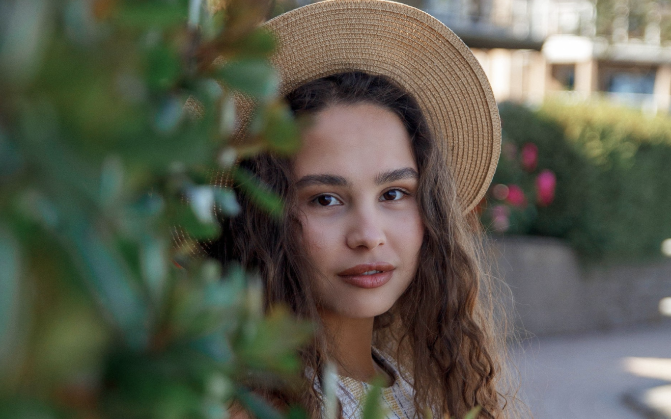 Anna Shynkarenko wearing a hat, half-hidden behind a bush and looking at the camera