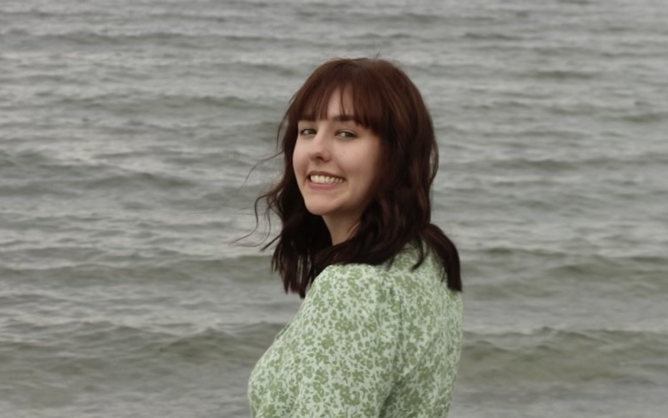 Paige Pierce standing in front of the ocean, wearing a green dress and turning back to smile at the camera