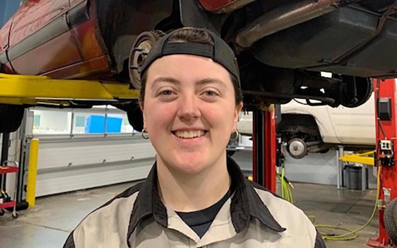 Trinity stands in front of a car raised up on a stand